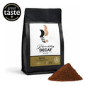 A matt black, pack of Brazilian, Raw Bean Definitely Decaf, on a white background with a Great Taste Award badge and coffee grounds. The label shows the Swiss Water Process certification. Tasting Notes: smooth, malty, cashew nut, soft caramel finish. Medium Roast. Strength 2, 200g.