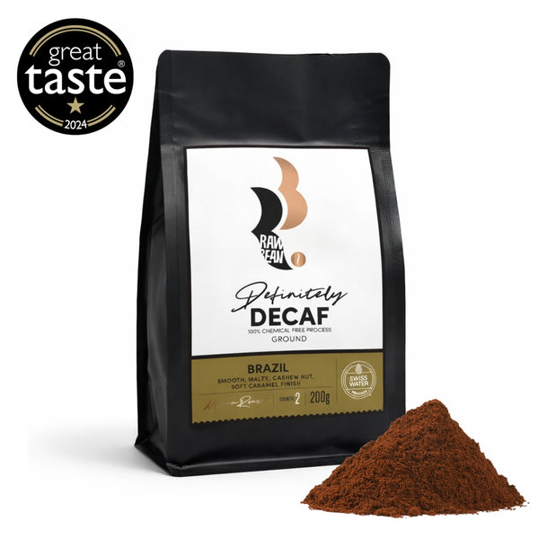 A matt black, pack of Brazilian, Raw Bean Definitely Decaf, on a white background with a Great Taste Award badge and coffee grounds. The label shows the Swiss Water Process certification. Tasting Notes: smooth, malty, cashew nut, soft caramel finish. Medium Roast. Strength 2, 200g.