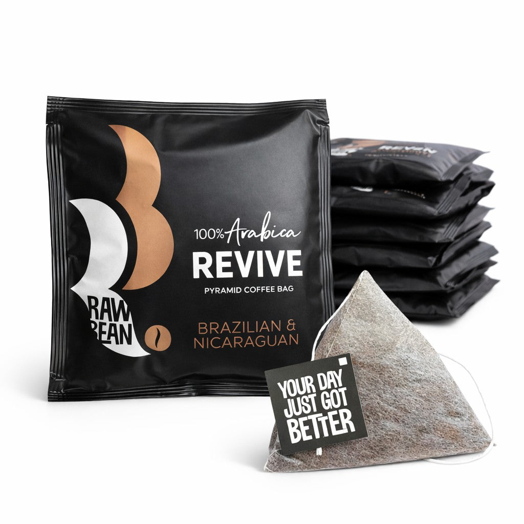 The foreground shows a square, matt black, envelope with a white and gold Raw Bean steam logo. The pack reads: 100% Arabica Revive Pyramid Coffee Bag, Brazilian & Nicaraguan. In front of the envelope is a pyramid coffee bag with a string and tag reading: Your Day Just Got Better. In the background is a pile of more envelopes.