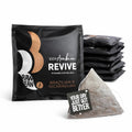 The foreground shows a square, matt black, envelope with a white and gold Raw Bean steam logo. The pack reads: 100% Arabica Revive Pyramid Coffee Bag, Brazilian & Nicaraguan. In front of the envelope is a pyramid coffee bag with a string and tag reading: Your Day Just Got Better. In the background is a pile of more envelopes.