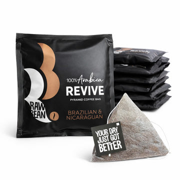 The foreground shows a square, matt black, envelope with a white and gold Raw Bean steam logo. The pack reads: 100% Arabica Revive Pyramid Coffee Bag, Brazilian & Nicaraguan. In front of the envelope is a pyramid coffee bag with a string and tag reading: Your Day Just Got Better. In the background is a pile of more envelopes.