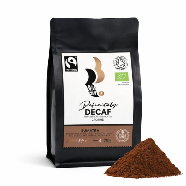 A matt black, pack of Sumatran, Raw Bean Definitely Decaf, on a white background with coffee grounds. The label shows certifications for Fairtrade, Organic and the 100% chemical free Swiss Water Process. Tasting Notes: Smokey molasses, spice, tropical fruits, full bodied and intense, bold finish. Medium Roast, Strength 4, 200g.