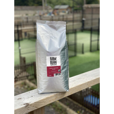 Raw Bean Unpacked Dark & Delicious part of the Waitrose Unpacked trial, pack shot on wooden ledge with Padel court in the background 