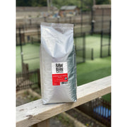 Raw Bean Unpacked Honduran Cafe Capucas part of the Waitrose Unpacked trial, pack shot on wooden ledge with Padel court in the background 