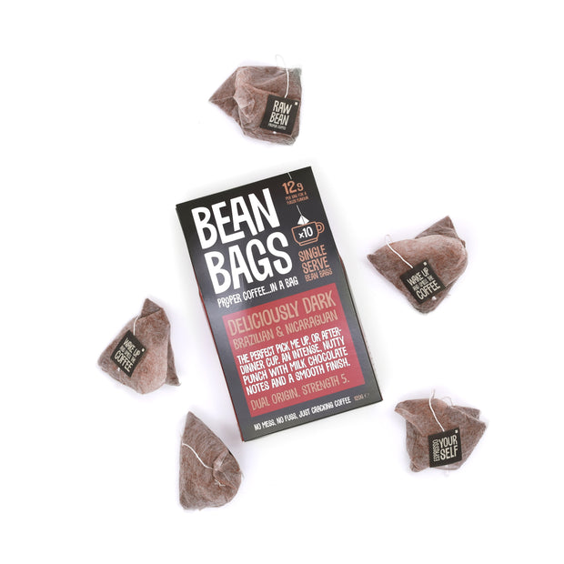 A pack of ten loose Deliciously Dark "Bean Bags", Proper Coffee... in a Bag. The pack is on a white background surrounded by pyramid coffee bags each with string and tag and containing 12g of coffee. The pack front describes the Brazilian & Nicaraguan Arabica blend: An intense nutty punch with milk chocolate notes and a smooth finish. Strength 5. Brought to you by Raw Bean.